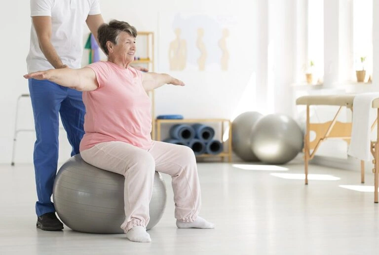 An elderly woman being assisted by a specialist while sitting on a medical ball, doing exercises.