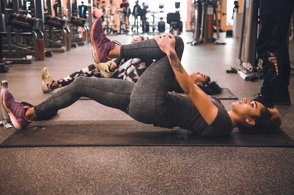 Two women lying on a gym floor and stretching their left legs with a person standing and observing them.