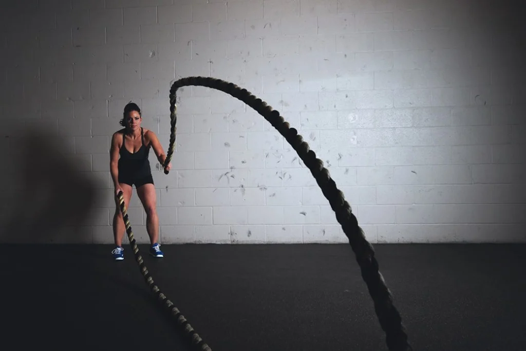 A muscular woman exercising with heavy long ropes in an empty room with tarmac floor.