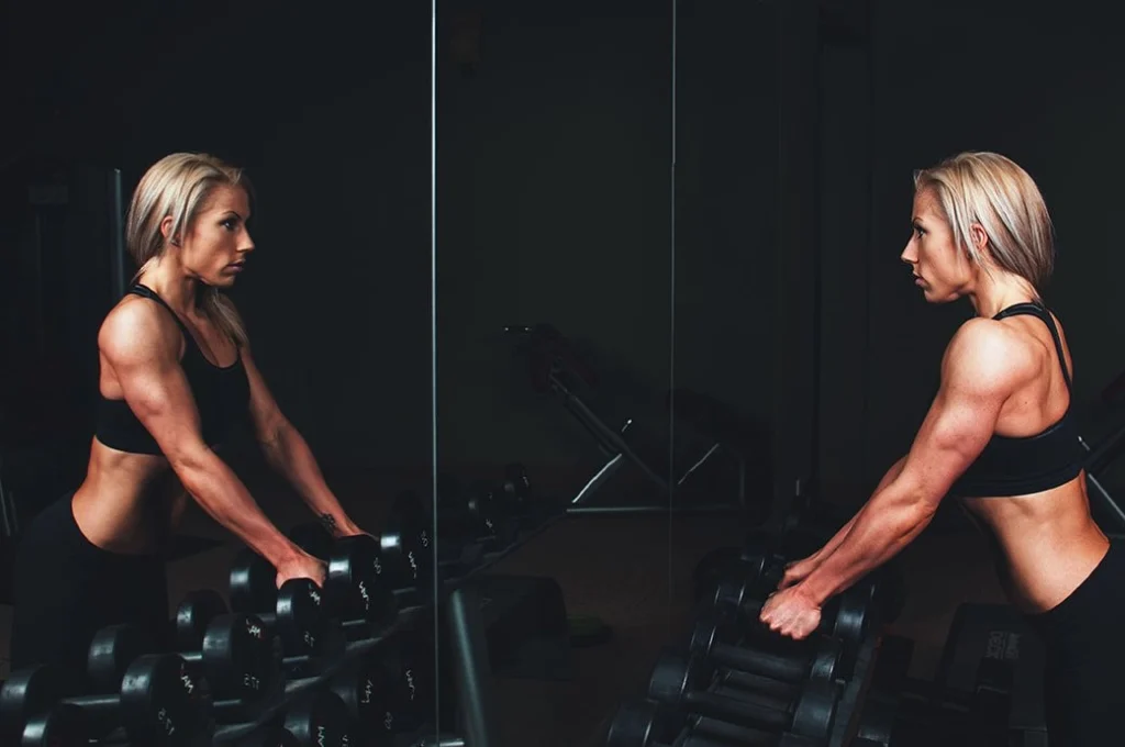 A muscular woman looking on the mirror of a gym while getting ready to pick up a pair of weights.