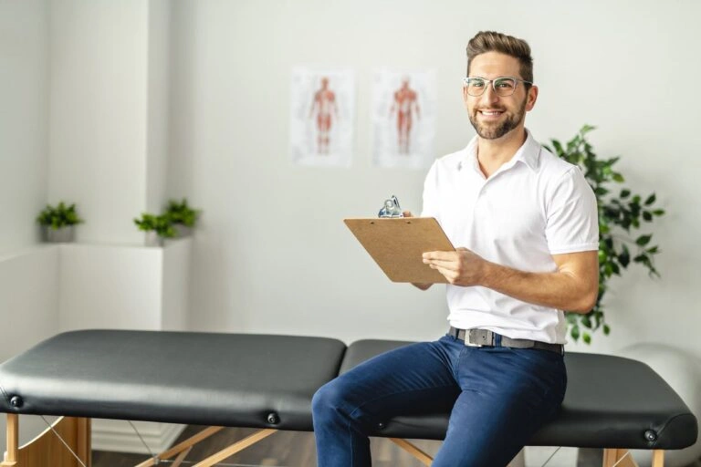 A happy chiropractor sitting on a massage table holding a notepad.