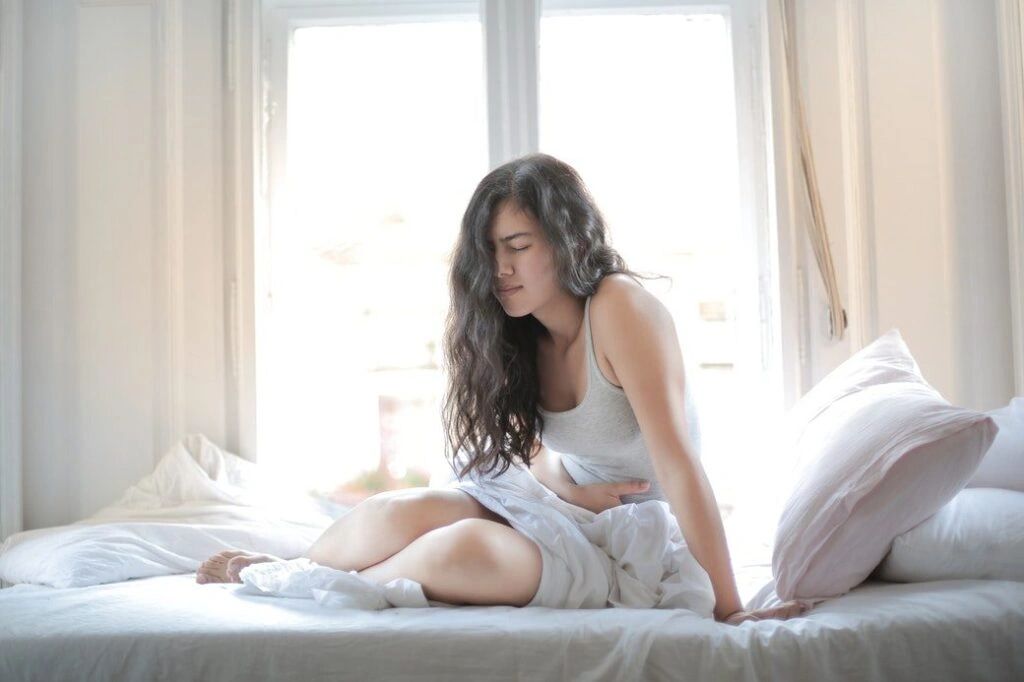 A young woman sitting on her bed looking to be in pain while touching her lower belly.