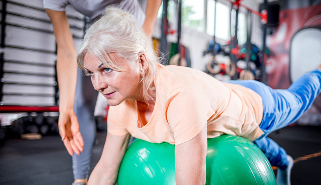 A woman doing exercises on a medical ball with the supervision of a specialist.