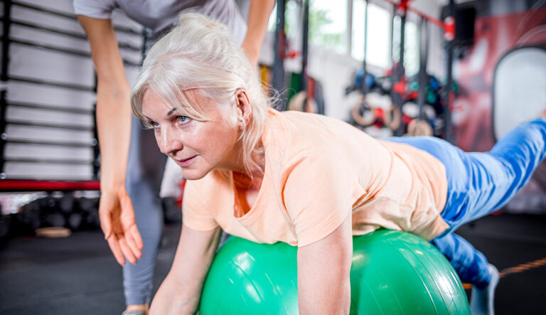 A woman doing exercises on a medical ball with the supervision of a specialist.