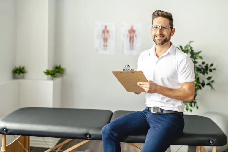 A man sitting on a massage table with a notepad, smiling.