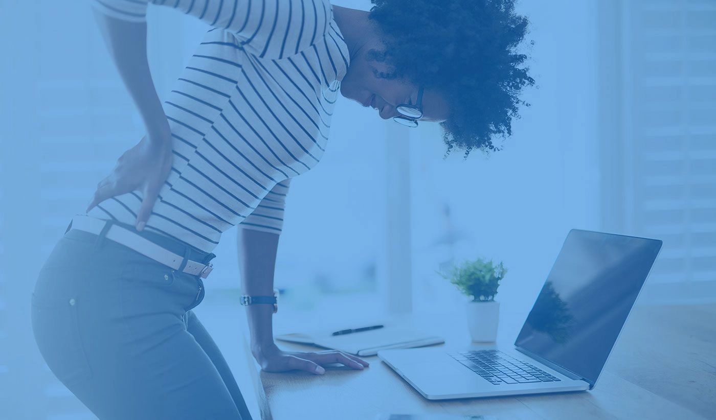 A woman touching her lower back and trying to stand in front o her desk.