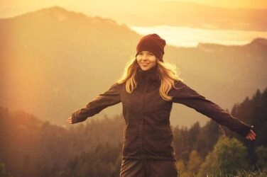 A girl with her arms open, in front of a distant mountain and forest scenery, looking happy.