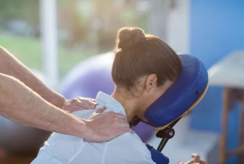 A woman lying facedown on a massage chair with her head supported that is being treated by a chiropractor on her neck.